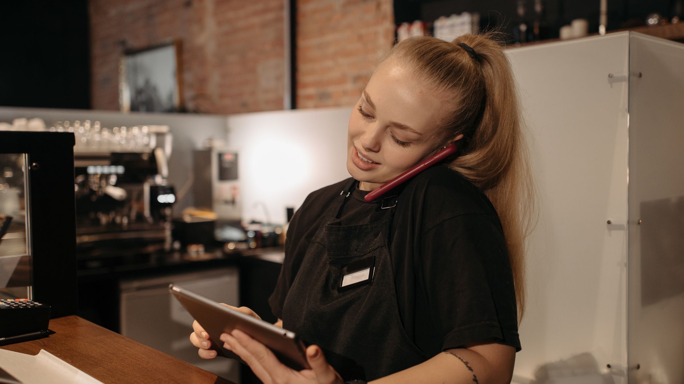 A restaurant manager behind the counter checking a tablet while on the phone, juggling the restaurant management tools that run a shift